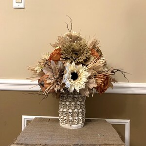 Puede incluir: Un arreglo floral de otoño en un jarrón de cerámica blanco con un diseño de calavera. El jarrón está lleno de flores secas en tonos de marrón, naranja y blanco.