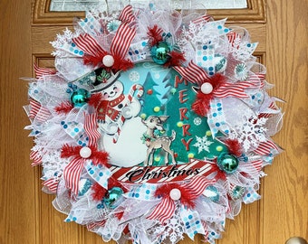 May include: A Christmas wreath featuring a snowman and reindeer design. The wreath is decorated with red and white striped ribbon, white mesh, and blue and white snowflake baubles. The wreath also includes the words "Merry Christmas".