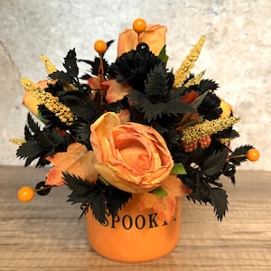 Może przedstawiać: Aranżacja kwiatowa na Halloween w pomarańczowym ceramicznym wazonie. Na wazonie napis "SPOOK". Aranżacja zawiera pomarańczowe róże, czarne liście i żółte akcenty, tworząc sezonową ekspozycję.