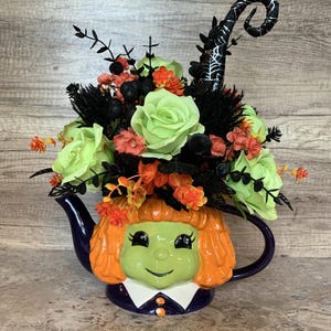 Könnte beinhalten: Eine Halloween-Teekanne mit einem Hexengesicht-Design. Die Teekanne ist mit grünen Rosen, orangefarbenen und schwarzen floralen Akzenten und einem schwarzen Hexenhut gefüllt. Die Teekanne hat einen lila Griff und Ausguss.