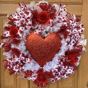 以下が含まれることがあります： 白と赤のリボン、赤いバラ、白いモールで飾られた、赤いグリッターハートのリース。