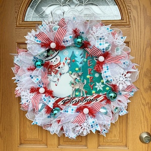 Puede incluir: Una corona de Navidad con un diseño de muñeco de nieve y reno. La corona está decorada con cinta a rayas rojas y blancas, malla blanca y adornos de copo de nieve azul y blanco. La corona también incluye las palabras "Feliz Navidad".