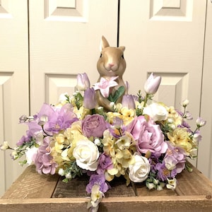 Puede incluir: Una figurita de conejo de cerámica que sostiene una flor se encuentra en el centro de un arreglo floral. El arreglo presenta una variedad de flores moradas, blancas y amarillas, incluyendo rosas, hortensias y tulipanes.