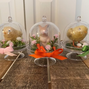 May include: Three clear glass cloche domes with a base, each containing a different Easter-themed figurine. The first dome contains a yellow chick, the second contains a pink bunny, and the third contains a yellow chick. Each dome has a ribbon tied around the base: pink, orange, and green.