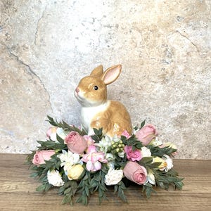 Könnte beinhalten: Eine Keramik-Hasenfigur sitzt auf einem Bett aus künstlichen rosa, weißen und gelben Blumen. Die Blumen sind kreisförmig angeordnet und von grünen Blättern umgeben.