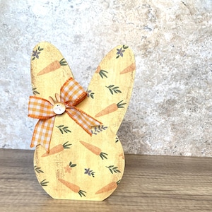 May include: A yellow wooden Easter bunny cutout with a carrot and leaf pattern. The bunny has a large orange and white gingham bow and a small brown button for a nose.