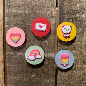 Op de afbeelding: Vijf ronde houten magneten met verschillende ontwerpen. De magneten zijn roze, rood, geel, groen en blauw. De ontwerpen omvatten een regenboog, een hart, een pandabeer, een ijslolly en een envelop met een hart.