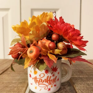May include: A white ceramic mug with a "thankful" design featuring orange and red fall leaves and berries. The mug is filled with a fall floral arrangement with orange, red, and yellow flowers.