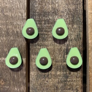 May include: Six green avocado-shaped pins with brown centres. The pins are arranged in a semi-circle on a wooden surface.