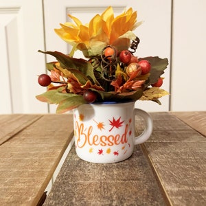 May include: A white ceramic mug with a blue rim and the word "Blessed" in orange lettering surrounded by small orange and red maple leaves. The mug is filled with a fall-themed floral arrangement featuring a yellow sunflower, red berries, and brown and green leaves.