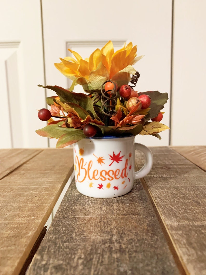May include: A white ceramic mug with a blue rim and the word "Blessed" in orange lettering surrounded by small orange and red maple leaves. The mug is filled with a fall-themed floral arrangement featuring a yellow sunflower, red berries, and brown and green leaves.