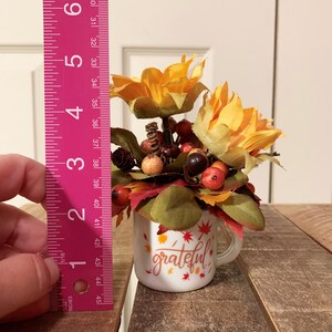 May include: A white ceramic mug with a handle, decorated with fall-colored leaves and the word "grateful". The mug is filled with a small arrangement of faux fall flowers, including yellow sunflowers, red berries, and brown pinecones.