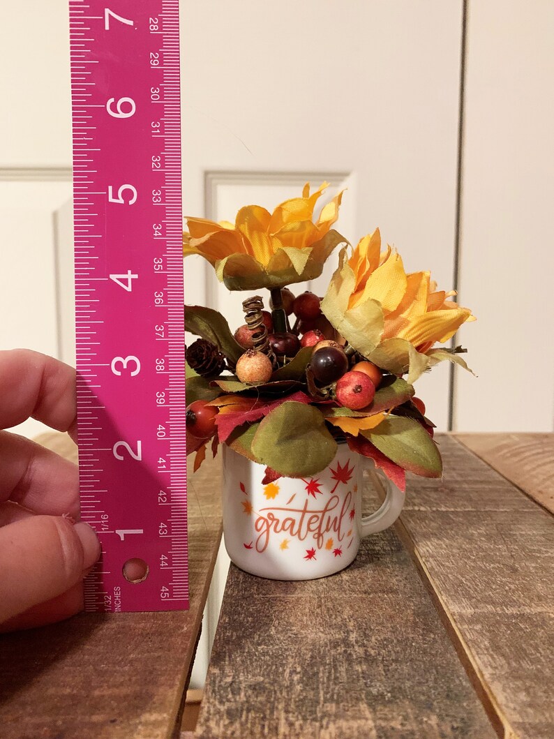May include: A white ceramic mug with a handle, decorated with fall-colored leaves and the word "grateful". The mug is filled with a small arrangement of faux fall flowers, including yellow sunflowers, red berries, and brown pinecones.