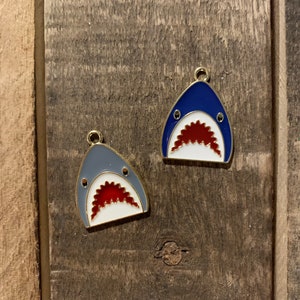 May include: Two metal shark charms with gold accents. One charm is blue with a white mouth and red teeth. The other charm is gray with a white mouth and red teeth.