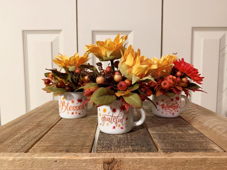 May include: Three white ceramic mugs with fall-themed designs. Each mug has a different word: "Blessed", "Grateful", and "Thankful". The mugs are filled with a floral arrangement of yellow sunflowers, red berries, and fall leaves.