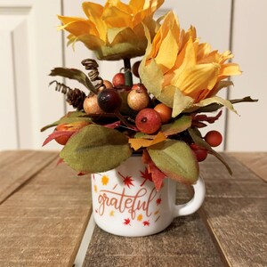 May include: A white ceramic mug with the word "grateful" written in orange and brown script. The mug is filled with a fall-themed arrangement of faux sunflowers, berries, and leaves.