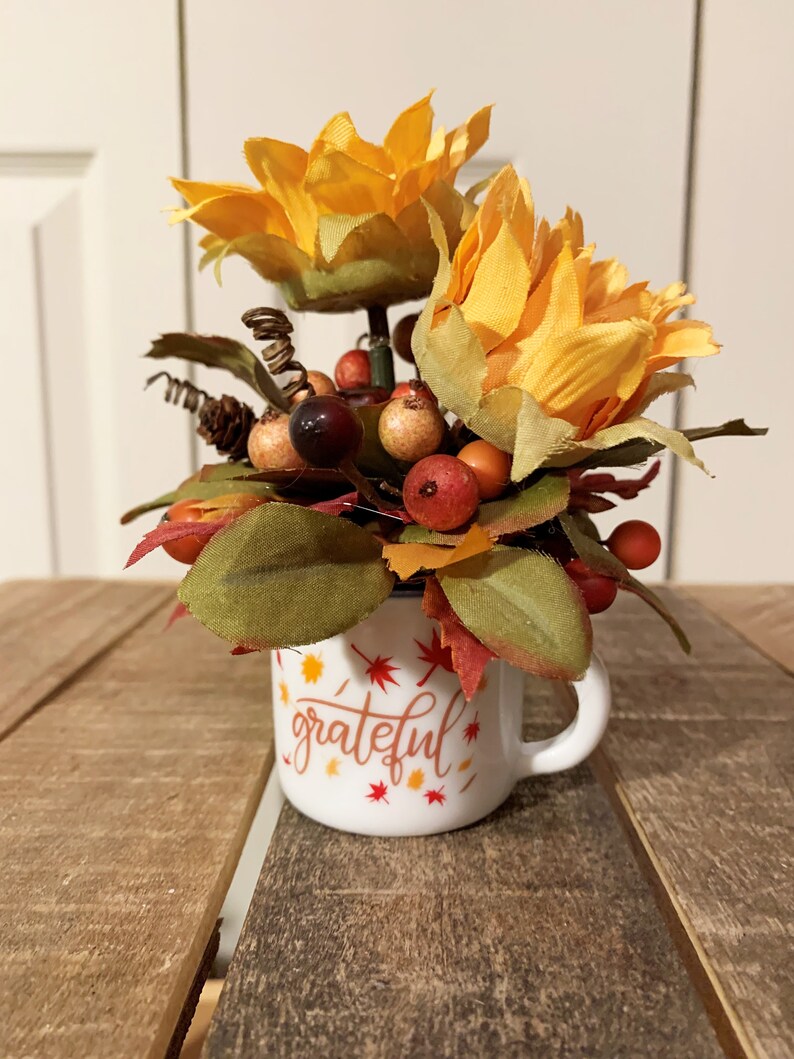 May include: A white ceramic mug with the word "grateful" written in orange and brown script. The mug is filled with a fall-themed arrangement of faux sunflowers, berries, and leaves.
