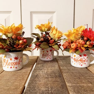 May include: Three white ceramic mugs with handles, each decorated with fall-themed designs and the words "Blessed", "Grateful", and "Thankful". Each mug is filled with a small arrangement of faux sunflowers, berries, and fall leaves.