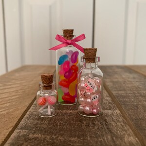 Könnte beinhalten: Drei kleine Glasgefäße mit Korkdeckeln gefüllt mit bunten Süßigkeiten, rosa Perlen mit Hasenanhängern und rosa und weißen Lutschern.