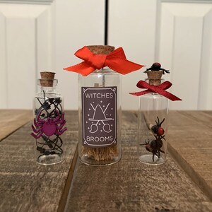 May include: Three small glass bottles with cork stoppers and red ribbon bows. The bottles contain various Halloween-themed items, including a spider, a broom, and a small red bug. The middle bottle has a label that reads "Witches Brooms".