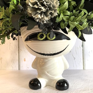 May include: A white ceramic mummy-shaped planter featuring black painted eyes with green irises and a wide grin. The planter is filled with a dense arrangement of dark green and black artificial foliage, including a single grey flower and a small lime green spider.