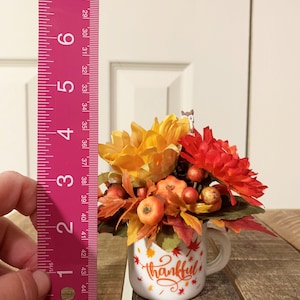 May include: A white ceramic mug with a handle, decorated with red and orange fall leaves and the word "thankful". The mug is filled with a fall floral arrangement featuring orange and red flowers, small pumpkins, and berries.
