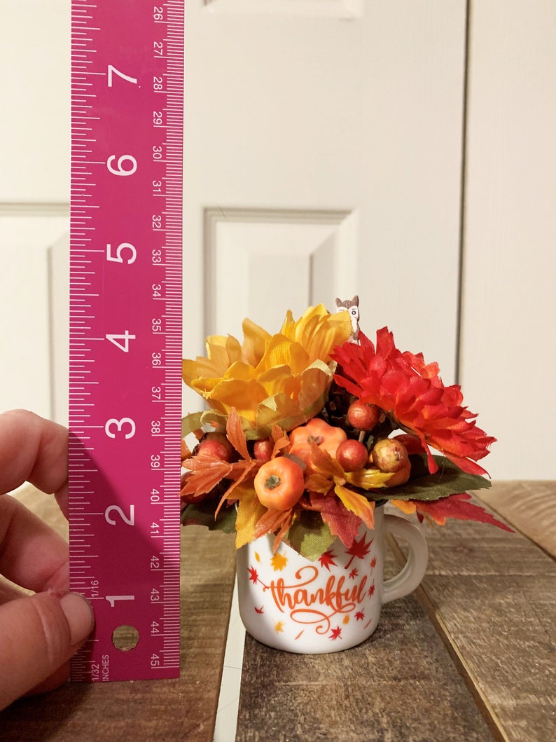 May include: A white ceramic mug with a handle, decorated with red and orange fall leaves and the word "thankful". The mug is filled with a fall floral arrangement featuring orange and red flowers, small pumpkins, and berries.