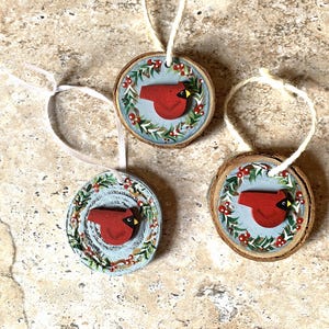 May include: Three round wooden ornaments with a red cardinal design. Each ornament features a red cardinal on a light blue background, surrounded by a wreath of green leaves and red berries. White string is attached for hanging.