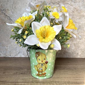 May include: A green metal bucket with white polka dots and a cartoon chick holding a basket of Easter eggs. The bucket is filled with a bouquet of white and yellow daffodils.