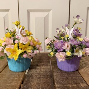 May include: Two Easter baskets filled with colorful flowers and a white bunny figurine. The basket on the left is blue and the basket on the right is purple.