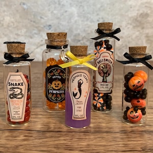 Puede incluir: Cinco botellas de vidrio decorativas con tapones de corcho, llenas de caramelos coloridos y etiquetadas con texto temático de Halloween. Las botellas están adornadas con lazos negros y cintas amarillas.