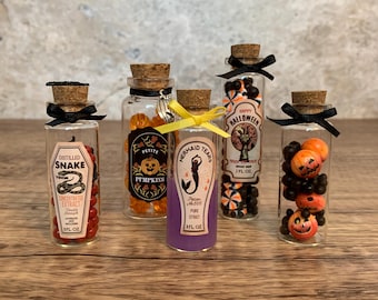 Puede incluir: Cinco botellas de vidrio decorativas con tapones de corcho, llenas de caramelos coloridos y etiquetadas con texto temático de Halloween. Las botellas están adornadas con lazos negros y cintas amarillas.