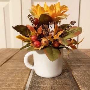 May include: A white ceramic mug filled with a fall-themed floral arrangement. The arrangement features a yellow sunflower, red berries, and brown leaves. A small wooden squirrel figurine is nestled in the leaves.