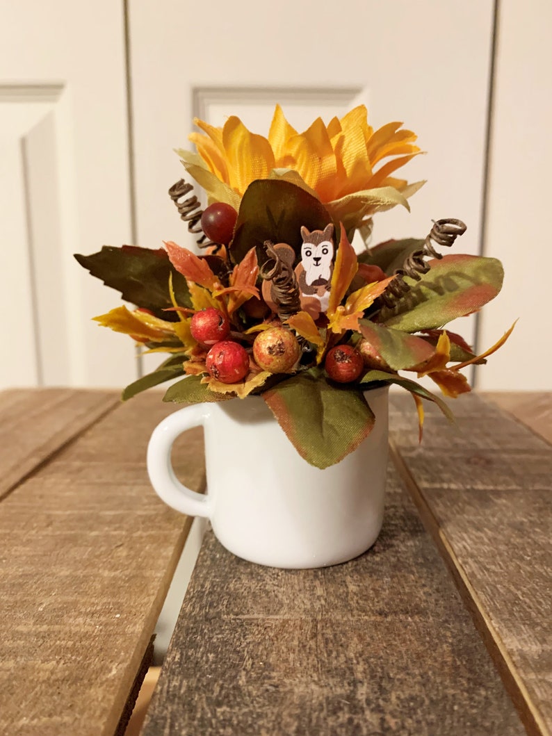 May include: A white ceramic mug filled with a fall-themed floral arrangement. The arrangement features a yellow sunflower, red berries, and brown leaves. A small wooden squirrel figurine is nestled in the leaves.