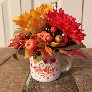 May include: A white ceramic mug with the word "thankful" written in red and orange lettering. The mug is filled with a fall-themed arrangement of orange and red flowers, green leaves, and small red berries.