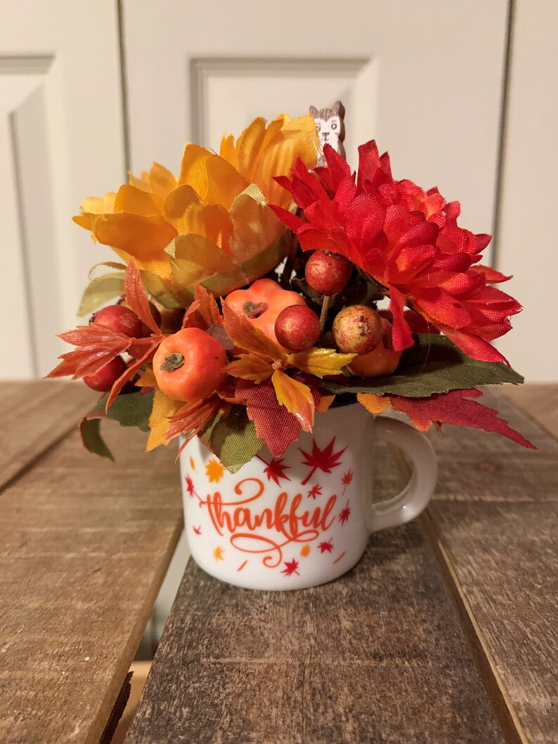 May include: A white ceramic mug with the word "thankful" written in red and orange lettering. The mug is filled with a fall-themed arrangement of orange and red flowers, green leaves, and small red berries.