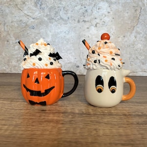 May include: Two Halloween-themed mugs. One is orange and shaped like a jack-o'-lantern with a black handle. The other is white and shaped like a ghost with an orange handle. Both mugs are topped with whipped cream and decorative sprinkles.
