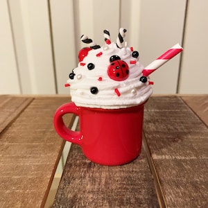 Peut inclure: Une tasse en céramique rouge avec un bord blanc, remplie de glaçage blanc, de pépites rouges et noires, de cannes en sucre noir et blanc, de perles blanches et une coccinelle rouge sur le dessus. Une paille rayée rouge et blanche sort du glaçage.
