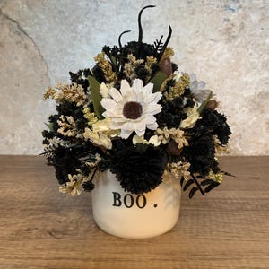 Puede incluir: Una maceta de cerámica blanca con la palabra "BOO" en letras negras, llena de un arreglo de flores artificiales negras, blancas y beige. El arreglo incluye una gran margarita blanca con un centro marrón.