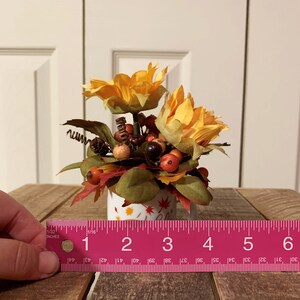May include: A small white ceramic mug with a fall-themed design, decorated with a bouquet of yellow sunflowers, red berries, and brown leaves. The mug is approximately 3 inches tall.