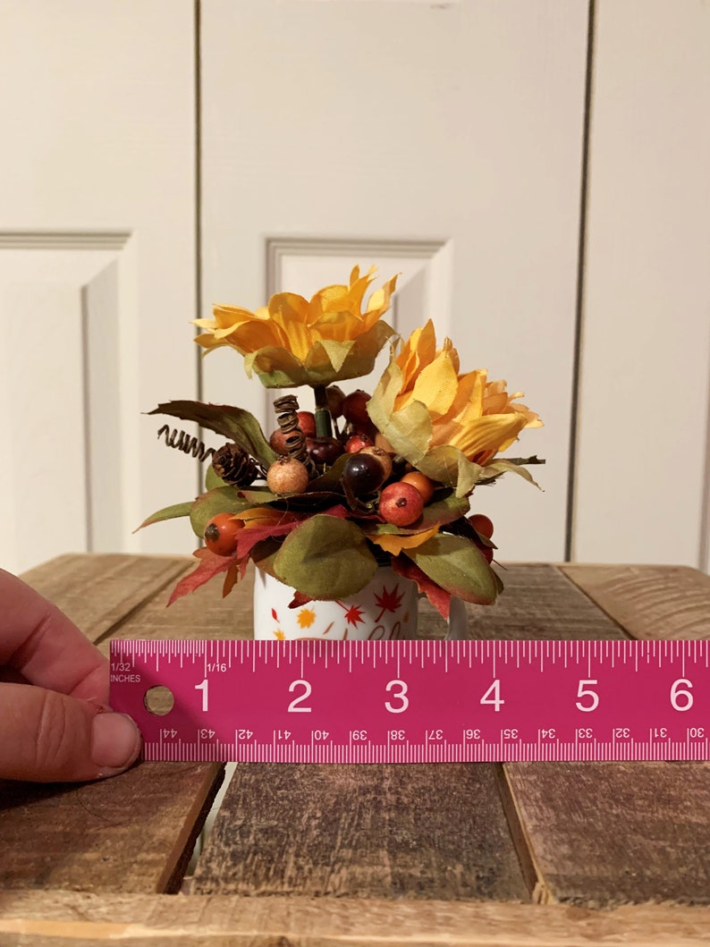 May include: A small white ceramic mug with a fall-themed design, decorated with a bouquet of yellow sunflowers, red berries, and brown leaves. The mug is approximately 3 inches tall.