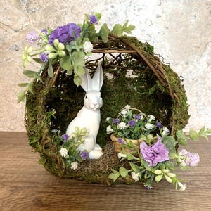 May include: A white ceramic bunny figurine sits inside a woven wreath decorated with moss, green leaves, and purple flowers. The wreath is a spring-themed decoration.