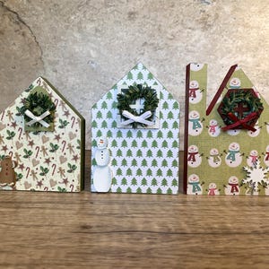 Op de afbeelding: Drie decoratieve houten huisjes met kerstpatronen. Elk huis heeft een kleine krans boven een raam. De huizen zijn versierd met peperkoekmannetjes, zuurstokken, sneeuwpoppen en kerstbomen. De huizen staan op een houten oppervlak.