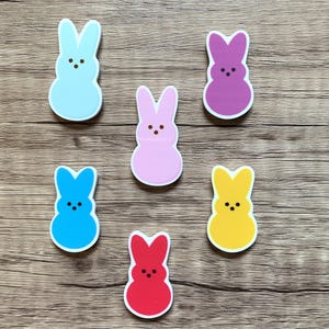 May include: Six colorful bunny-shaped marshmallow candies in various colors including blue, pink, purple, yellow, and red. The candies are arranged on a woodgrain surface.