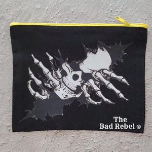 May include: Black canvas pouch with a yellow zip and trim. The design features a skull and skeletal hands breaking through a torn surface. The text "The Bad Rebel" is printed on the bottom right of the pouch.