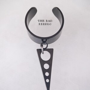 Peut inclure: Boucle d'oreille noire avec un pendentif triangulaire orné de plusieurs cercles découpés. La boucle d'oreille est gravée du texte "THE BAD REBELO".