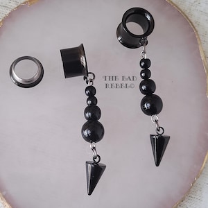 May include: Black ear gauges with dangling beaded details and a cone-shaped charm. The gauges are displayed on a white and pink surface. The text "THE BAD REBELO" is visible.