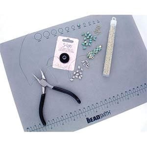 May include: A grey craft mat with a ruler and circle guides, holding beading supplies. Includes a package of S-Lon thread, pliers, turquoise and silver beads, and a tube of small, gold-coloured beads. The mat is branded with the name "BeadSmith."