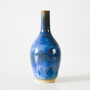 Op de afbeelding: Een keramische vaas met een smalle hals en een bolvormig lichaam, voorzien van een gespikkeld glazuur in tinten diep kobaltblauw, marineblauw en lichter azuurblauw. Het oppervlak heeft een glanzende afwerking met fijne donkere spikkels en een beige voet.