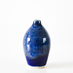 Op de afbeelding: Een keramische vaas met een smalle hals en een ronde vorm, voorzien van een diep kobaltblauw glazuur met een gevlekte, getextureerde afwerking die overgaat in een donkerdere, effen blauwe tint bij de basis, met een zichtbare lichte keramische rand.
